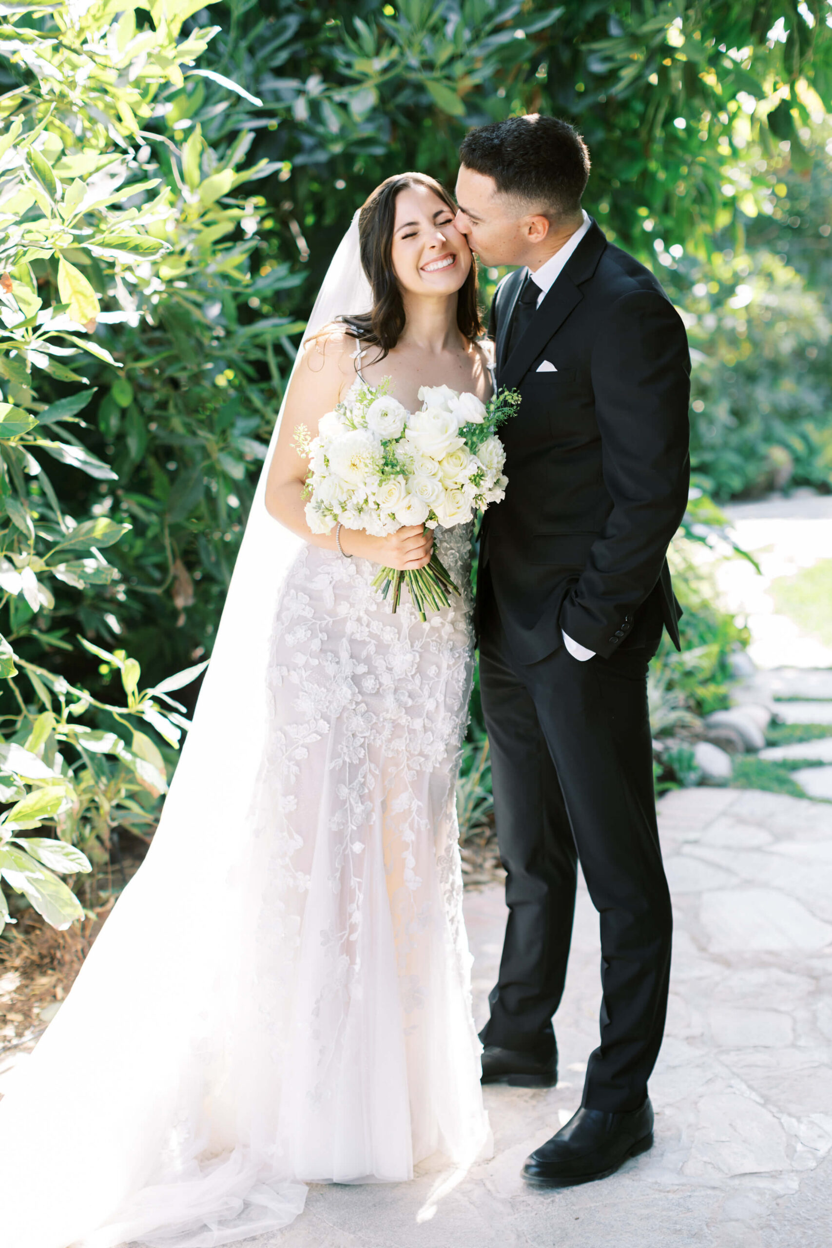A groom in a black suit kisses a smiling bride in a white wedding dress holding a bouquet of white flowers. They stand on a garden path surrounded by greenery and sunlight. Copyright - Lisa Leanne Photography, California