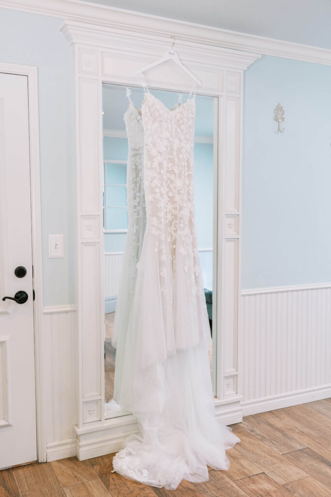 Gerry Ranch Bridal Suite - A white lace wedding dress with floral appliqué details hangs on a hanger in front of a large decorative mirror in a softly lit room with light blue walls and wood flooring. Copyright - Lisa Leanne Photography, California