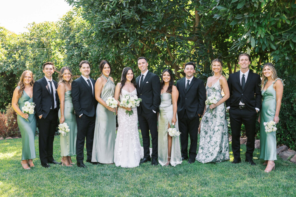 Gerry Ranch Wedding Bridal Party - A wedding party poses outdoors, with six women in light green dresses holding bouquets and five men in black suits standing beside the bride in a white gown and the groom in a black suit. Lush greenery is in the background. Copyright - Lisa Leanne Photography, California
