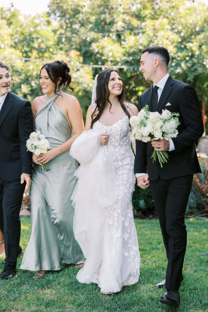 Gerry Ranch Wedding Bridal Party - A bride in a white lace gown and a groom in a black suit walk hand in hand outdoors, smiling. A bridesmaid in a green dress and another man in a suit walk beside them, all holding bouquets and celebrating. Copyright - Lisa Leanne Photography, California