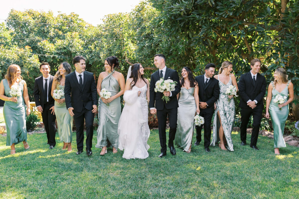 Gerry Ranch Wedding Bridal Party - A bride and groom walk outdoors with their wedding party. The bridesmaids wear light green dresses and the groomsmen wear black suits. Everyone is smiling and holding bouquets, surrounded by lush greenery. Copyright - Lisa Leanne Photography, California
