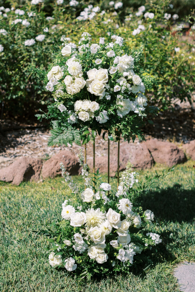 Gerry Ranch Wedding Ceremony - Two elegant floral arrangements of white roses and greenery are displayed outdoors on a grassy lawn, with a stone border and green bushes in the background. Copyright - Lisa Leanne Photography, California