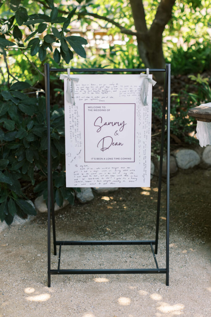 Gerry Ranch Wedding Ceremony - A wedding welcome sign for Sammy & Dean hangs on a black metal stand outdoors, surrounded by greenery. Handwritten notes are visible on the sign’s border. Sunlight filters through the trees onto the scene. Copyright - Lisa Leanne Photography, California