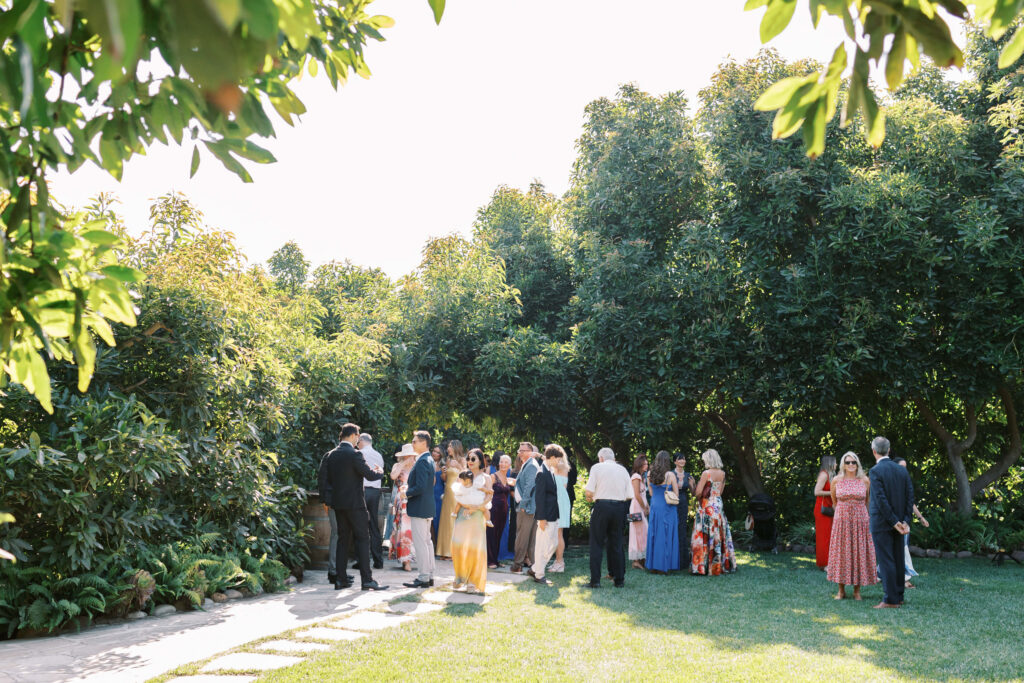 Gerry Ranch Wedding Ceremony - Guests people dressed in colorful, semi-formal attire gathers outdoors on a sunny day, standing on grass and near lush green trees, socializing before wedding ceremony. Copyright - Lisa Leanne Photography, California
