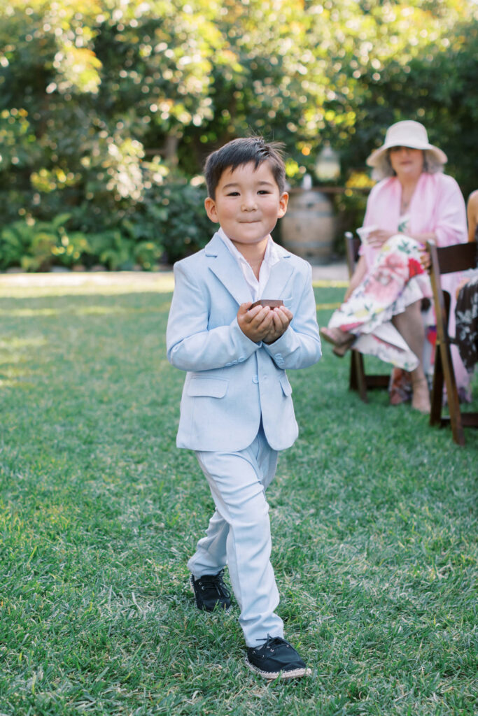 Gerry Ranch Wedding Ceremony - Ring bearer in a light blue suit walks on grass, holding something cupped in his hands. Copyright - Lisa Leanne Photography, California