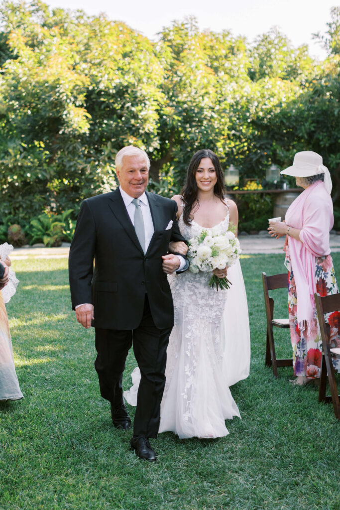 Gerry Ranch Wedding Ceremony - A bride in a white dress holding a bouquet walks arm-in-arm with an older man in a suit down a grassy aisle at an outdoor wedding, with guests seated nearby and greenery in the background. Copyright - Lisa Leanne Photography, California