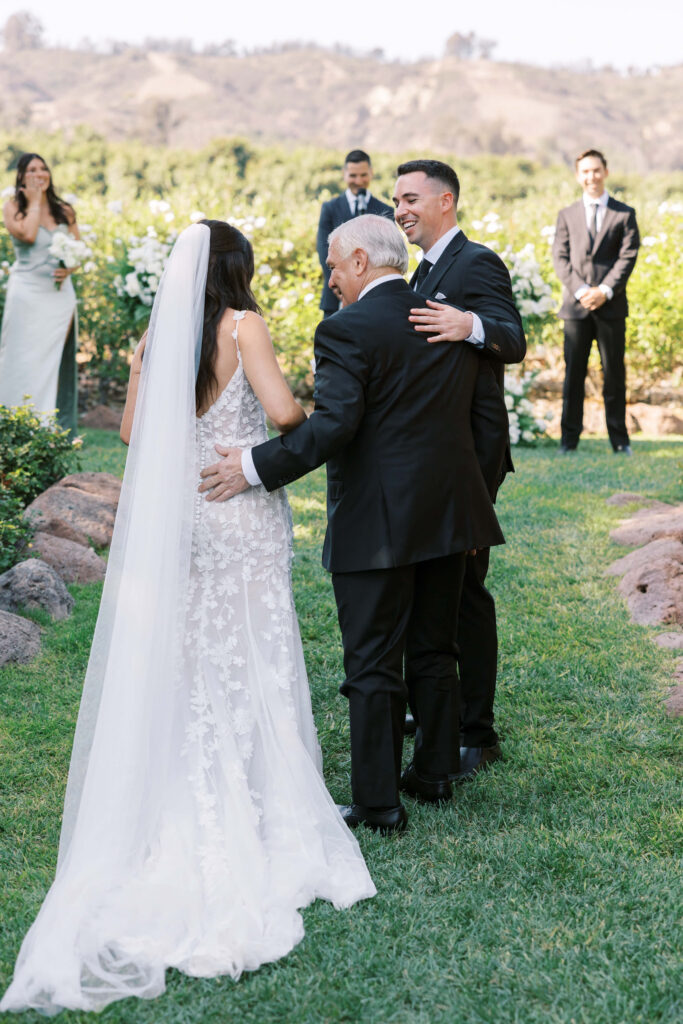 Gerry Ranch Wedding Ceremony - A bride in a white lace gown and veil is walked down an outdoor aisle by an older man, as a smiling groom greets them; bridesmaids and groomsmen stand in the background among greenery. Copyright - Lisa Leanne Photography, California