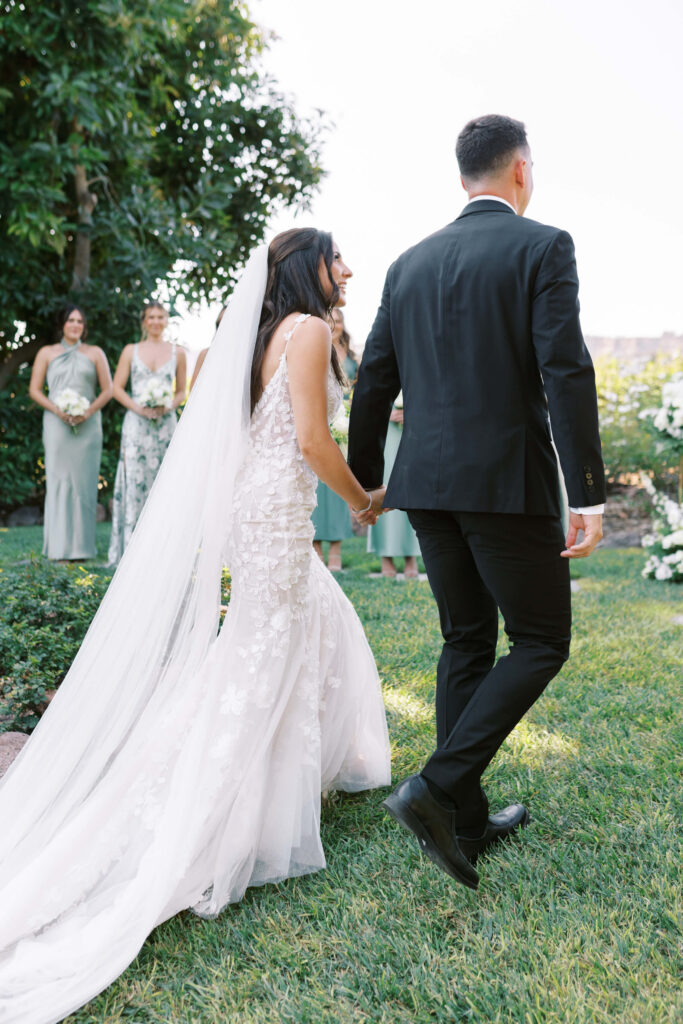 Gerry Ranch Wedding Ceremony - A bride in a white, floral wedding gown and veil holds hands with a groom in a black suit as they walk on grass outdoors. Bridesmaids in green dresses stand in the background, holding bouquets. Copyright - Lisa Leanne Photography, California