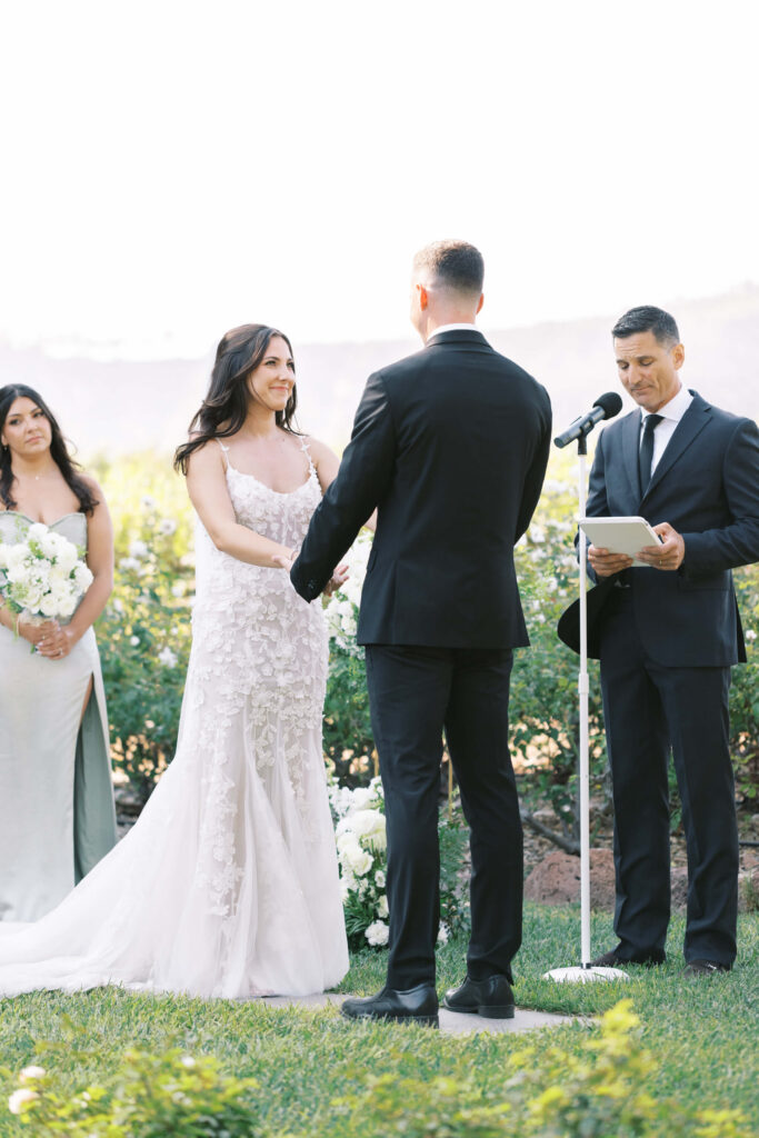 Gerry Ranch Wedding Ceremony - A bride and groom stand facing each other holding hands during an outdoor wedding ceremony. A bridesmaid in a light dress and a man in a suit reading from a book stand nearby. It is sunny with greenery in the background. Copyright - Lisa Leanne Photography, California