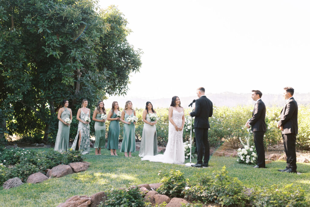 Gerry Ranch Wedding Ceremony - A bride and groom stand facing each other outdoors during their wedding ceremony. Bridesmaids in green dresses and groomsmen in black suits stand on either side, surrounded by greenery and flowers on a sunny day. Copyright - Lisa Leanne Photography, California
