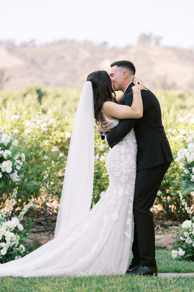 Gerry Ranch Wedding Ceremony - A bride in a white gown and veil kisses a groom in a black suit outdoors, surrounded by white flowers and greenery with hills in the background. Copyright - Lisa Leanne Photography, California