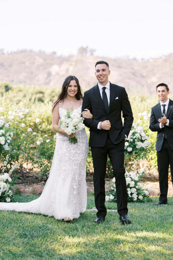 Gerry Ranch Wedding Ceremony - A bride in a white gown holding a bouquet walks arm-in-arm with a groom in a black suit outdoors, smiling, with greenery and white flowers in the background and a man clapping nearby. Copyright - Lisa Leanne Photography, California