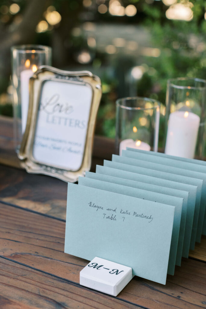 Gerry Ranch Reception Seating Chart Table - A stack of blue envelopes labeled with guests' names sits on a wooden table, sorted by an 