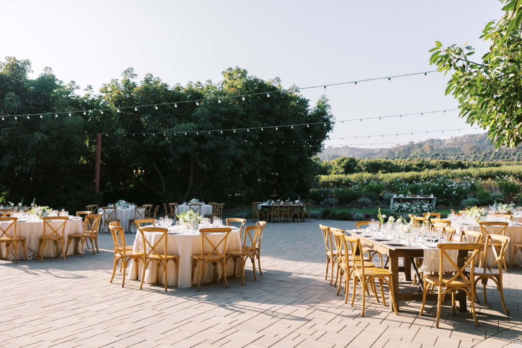 Gerry Ranch Reception Setup with round and rectangular tables covered in white linens, surrounded by wooden chairs, string lights overhead, and lush green trees in the background on a sunny day. Copyright - Lisa Leanne Photography, California
