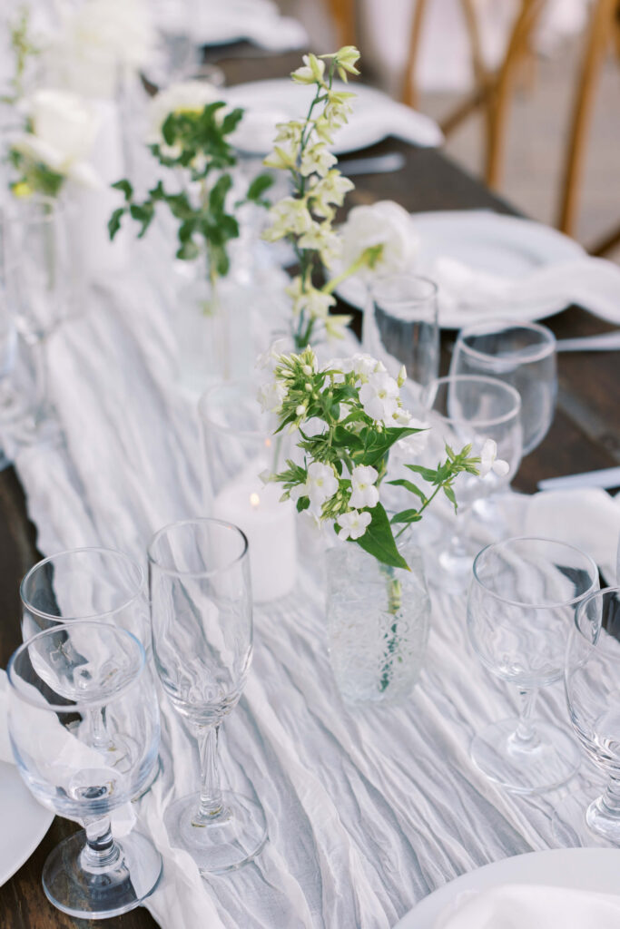 Gerry Ranch Reception Detail - A close-up of an elegant table setting with clear glassware, white plates, and white flowers in glass vases on a light, crinkled table runner, creating a fresh and sophisticated atmosphere. Copyright - Lisa Leanne Photography, California