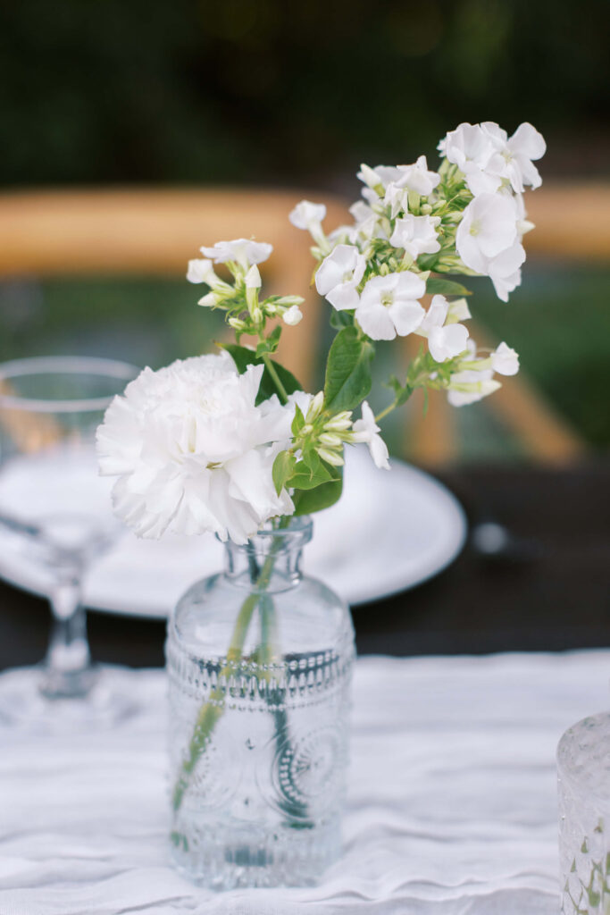 Gerry Ranch Reception Floral Detail - A small clear glass vase with white flowers sits on a table covered with a white cloth, with a blurred plate and glass in the background. Copyright - Lisa Leanne Photography, California
