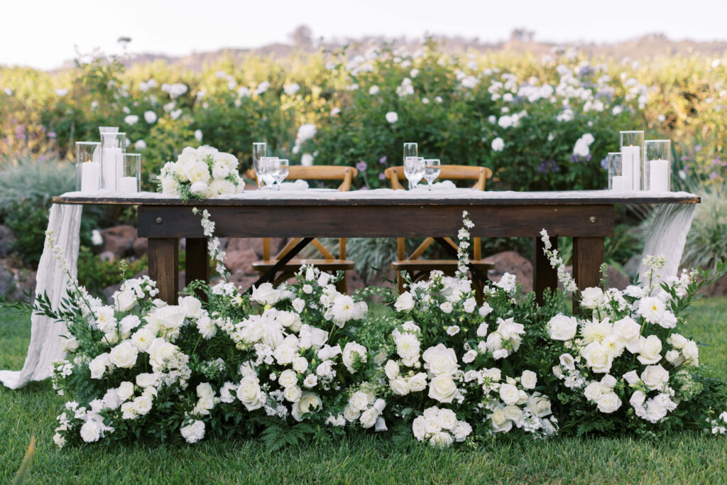 Gerry Ranch Reception Bride and Groom Table - A rustic wooden table is adorned with white flowers, glassware, and white candles in lanterns. Lush greenery and white roses surround the table, set outdoors amid blooming bushes and greenery. Copyright - Lisa Leanne Photography, California