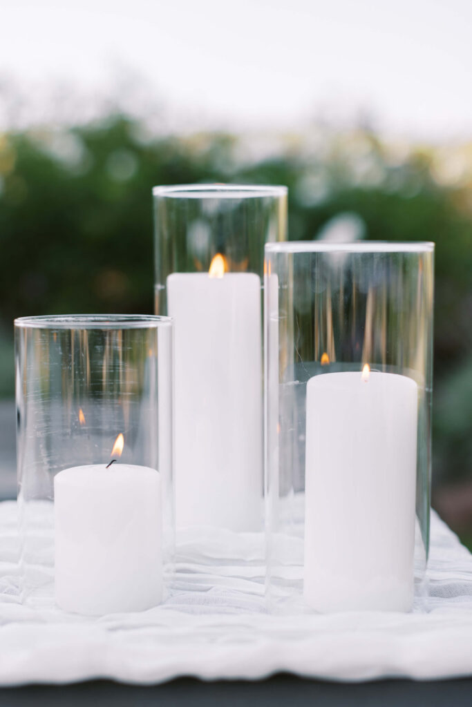 Gerry Ranch Reception Detail - Four white pillar candles, three enclosed in tall clear glass holders and one without a holder, are lit and placed on a white surface with greenery blurred in the background. Copyright - Lisa Leanne Photography, California