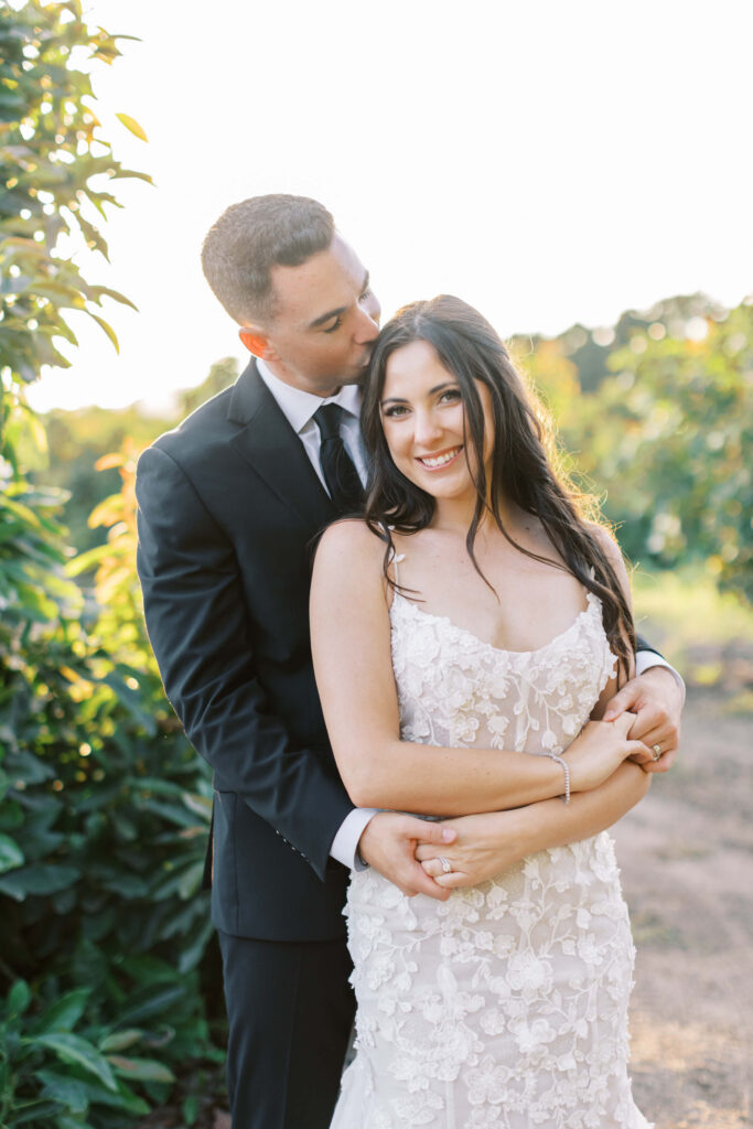 Golden Hour Bride and Groom Portrait at Gerry Ranch - A groom in a black suit gently embraces and kisses his smiling bride, who wears a white floral lace dress, as they stand outdoors surrounded by greenery in soft sunlight. Copyright - Lisa Leanne Photography, California