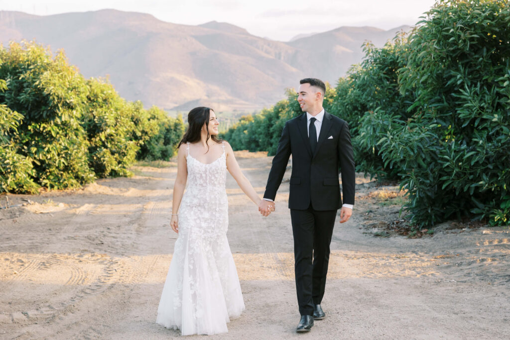 Golden Hour Bride and Groom Portrait at Gerry Ranch - A bride in a white dress and a groom in a black suit hold hands and walk on a dirt path between vineyards, with mountains in the background. Copyright - Lisa Leanne Photography, California