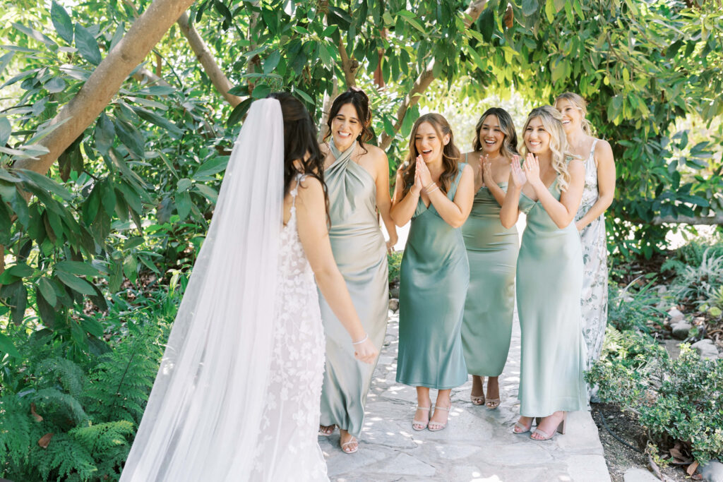 Bride first look with Bridesmaids at Gerry Ranch - A bride in a white dress and veil walks toward five bridesmaids in light green dresses, who smile and clap excitedly in a lush, green garden setting. Copyright - Lisa Leanne Photography, California