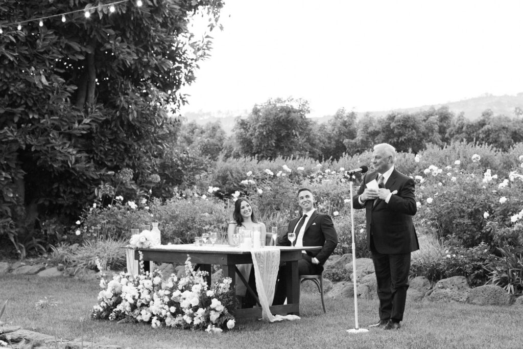 Gerry Ranch Wedding Reception - A man in a suit gives a speech at an outdoor wedding reception. A couple, seated at a decorated table with flowers, listen and smile. The setting is a garden with string lights and lush greenery. Copyright - Lisa Leanne Photography, California