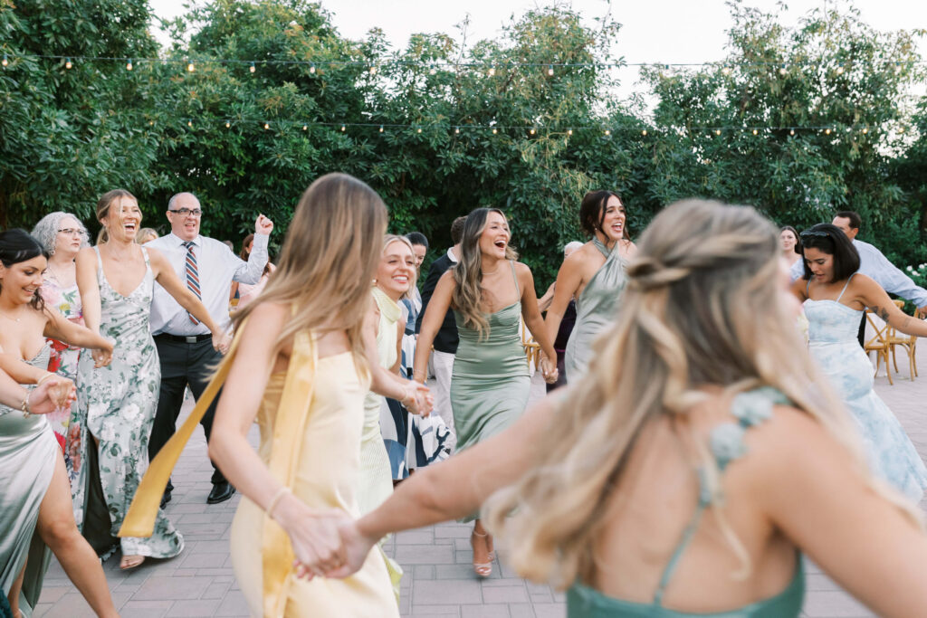 Gerry Ranch Wedding Reception Hora - A group of people in formal attire hold hands and dance in a circle outdoors, smiling and laughing under string lights, surrounded by greenery. Copyright - Lisa Leanne Photography, California