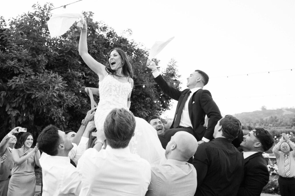 Gerry Ranch Wedding Reception Hora - A bride and groom are lifted on chairs by guests during an outdoor celebration. The bride waves a napkin and smiles joyfully, while people around them cheer and capture the moment with phones. Copyright - Lisa Leanne Photography, California