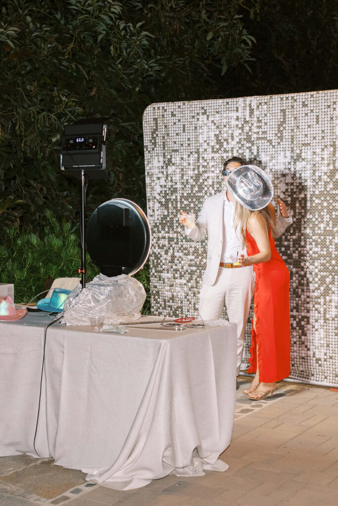 Gerry Ranch Wedding Reception Photo Booth - Two people in party attire pose together for a photo booth, wearing fun hats and kissing. They stand in front of a sparkly sequin backdrop, with a ring light and props on a table nearby. Copyright - Lisa Leanne Photography, California