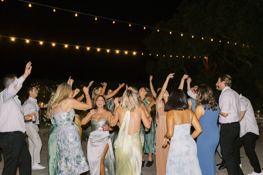 Gerry Ranch Wedding Reception Dance Floor - A group of people dressed in semi-formal attire dance joyfully outdoors at night under string lights, raising their arms and smiling. Copyright - Lisa Leanne Photography, California