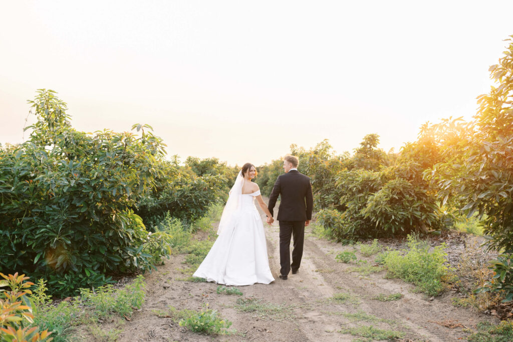 A bride and groom walk hand in hand down a dirt path between lush green bushes, with the sun setting in the background. The bride wears a white dress and veil, and the groom is dressed in a black suit. Copyright - Lisa Leanne Photography, California
