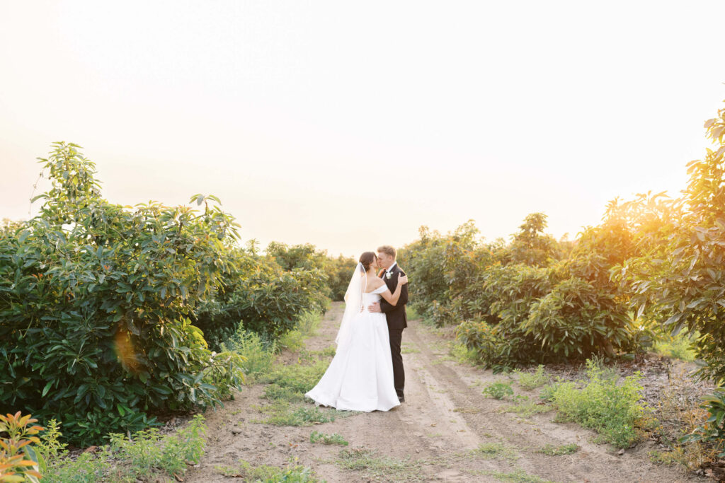 A bride and groom share a kiss on a dirt path surrounded by green plants or trees in a sunlit outdoor setting, with soft golden light illuminating the scene. Copyright - Lisa Leanne Photography, California