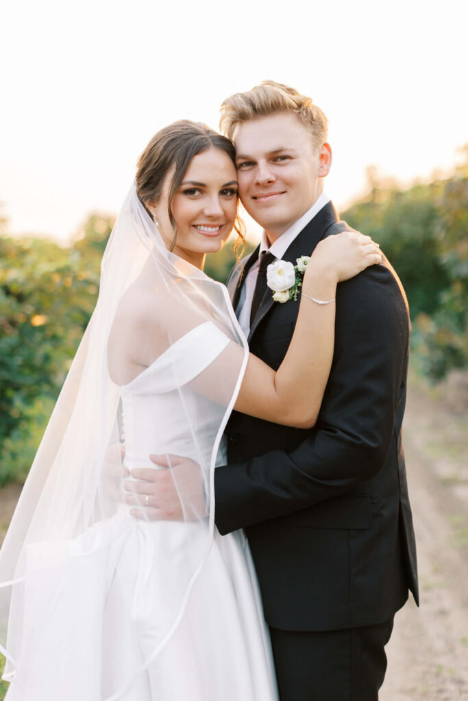 A bride in a white dress and veil embraces a groom in a black suit and white boutonnière outdoors, both smiling warmly at the camera. Greenery and soft sunlight are in the background. Copyright - Lisa Leanne Photography, California