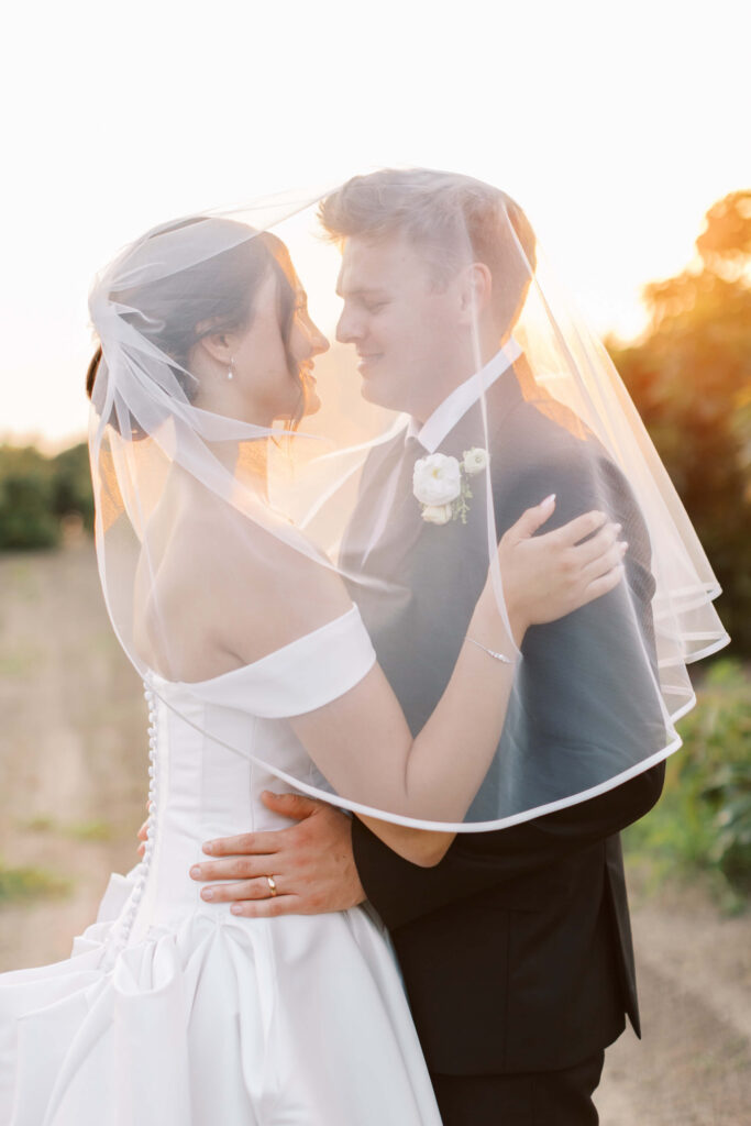 A bride and groom stand outdoors at sunset, smiling and embracing each other closely under the bride’s veil. The bride wears an off-shoulder white gown, and the groom is in a black suit with a white boutonnière. Copyright - Lisa Leanne Photography, California