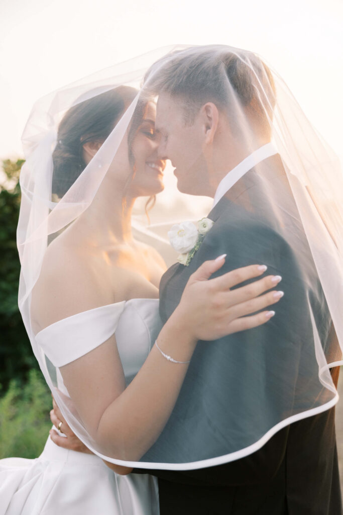 A bride and groom embrace closely, smiling under a sheer white veil outdoors. The bride wears an off-the-shoulder gown and the groom is in a dark suit with a white boutonniere. The sunlight gives a soft, romantic glow. Copyright - Lisa Leanne Photography, California