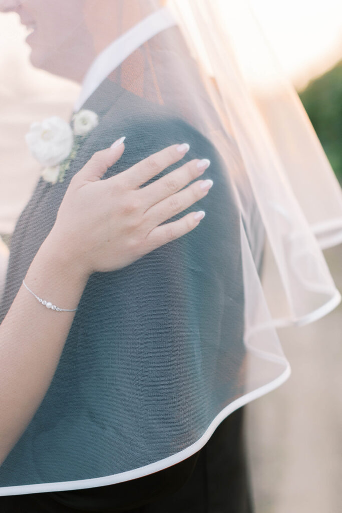 A close-up of a bride's hand gently resting on a groom's shoulder, both partially covered by a sheer wedding veil. The bride wears a delicate bracelet and has manicured nails; the groom has a boutonnière on his suit. Copyright - Lisa Leanne Photography, California