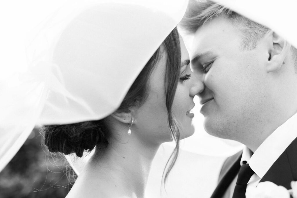 A black and white photo of a bride and groom, close together under a veil, gently touching noses and smiling with their eyes closed. Copyright - Lisa Leanne Photography, California