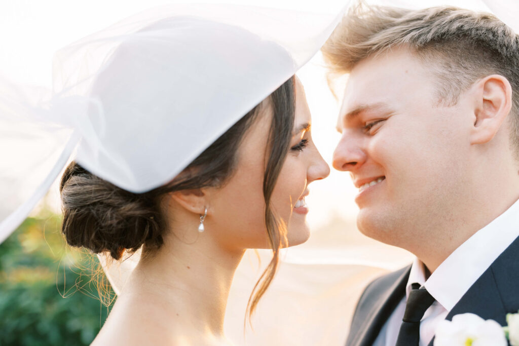 A bride and groom face each other closely, smiling with joy. The bride wears a veil and pearl earrings, and the groom is in a suit and tie. Soft sunlight creates a warm, romantic atmosphere. Copyright - Lisa Leanne Photography, California