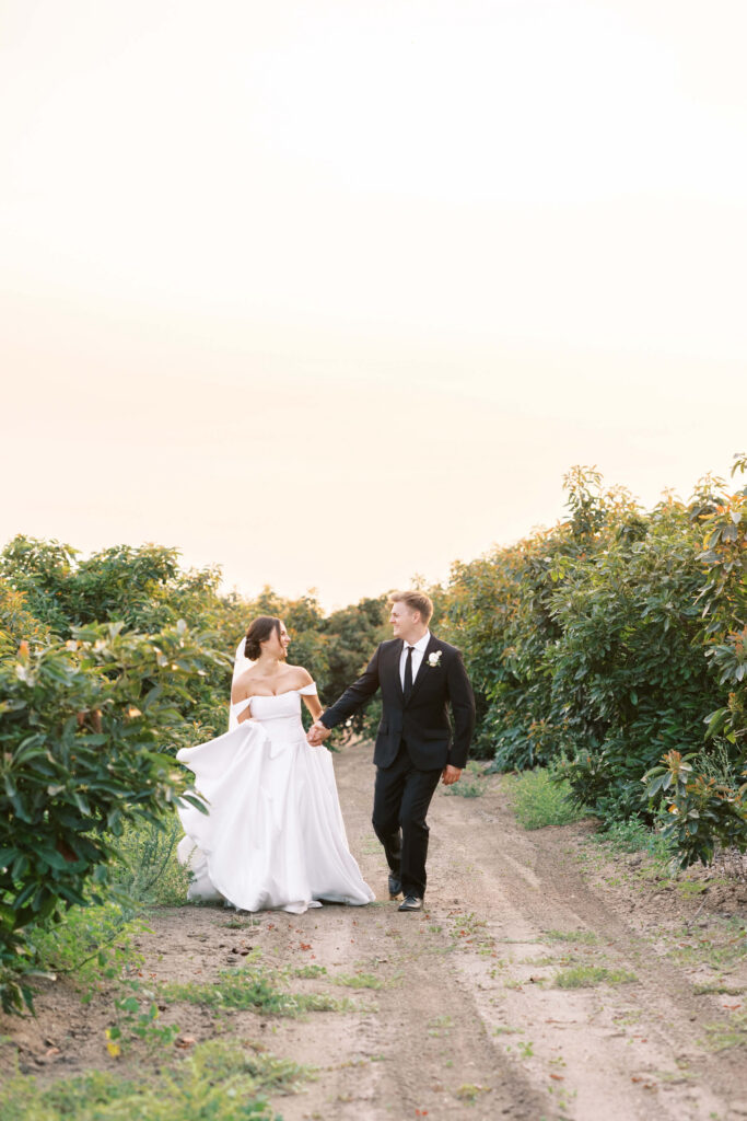 A bride in a white gown and a groom in a black suit walk hand in hand down a dirt path, surrounded by lush green bushes, under a soft, pastel sky. Copyright - Lisa Leanne Photography, California