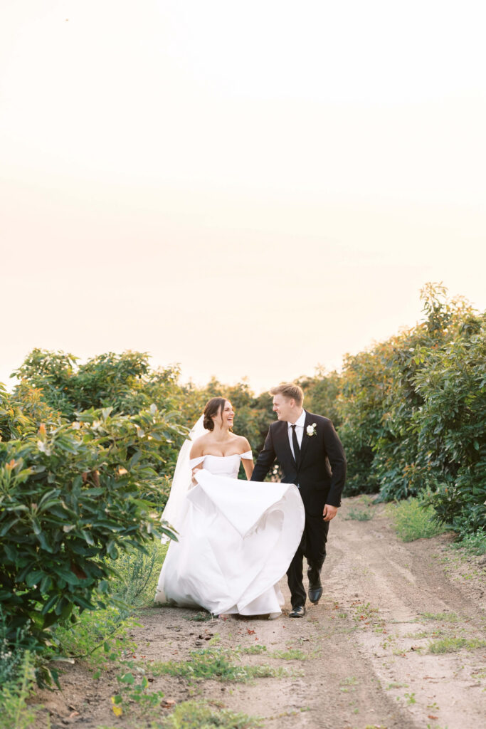 A bride in a white gown and veil walks hand in hand with a groom in a black suit and tie along a dirt path surrounded by green bushes, both smiling and looking at each other. Copyright - Lisa Leanne Photography, California