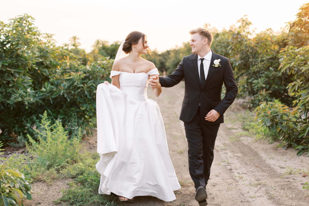 A bride in a white off-the-shoulder gown and a groom in a black suit walk hand-in-hand down a dirt path lined with greenery, smiling at each other on their wedding day. Copyright - Lisa Leanne Photography, California