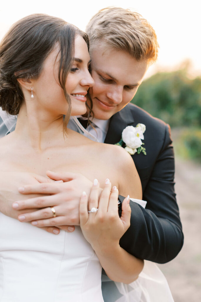 A bride and groom embrace outdoors. The bride smiles while showing her engagement ring, and the groom hugs her from behind, smiling with his eyes closed. Both look happy and dressed in wedding attire. Copyright - Lisa Leanne Photography, California