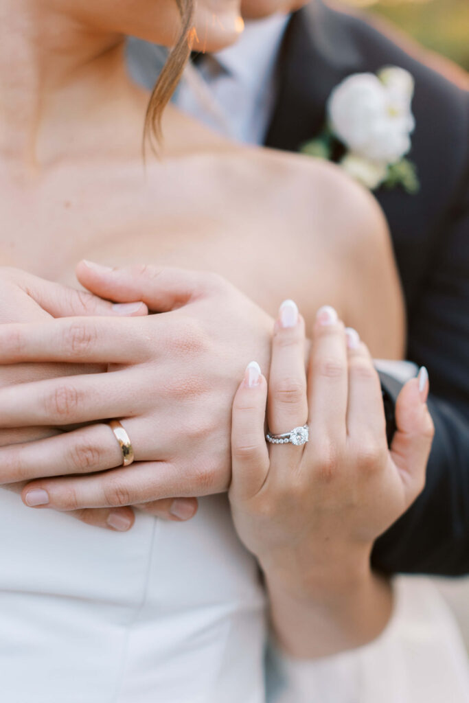 A close-up of a bride and groom embracing, showing their hands with wedding rings. The bride has a diamond engagement ring and French-manicured nails. The groom wears a gold band and a suit with a white boutonniere. Copyright - Lisa Leanne Photography, California