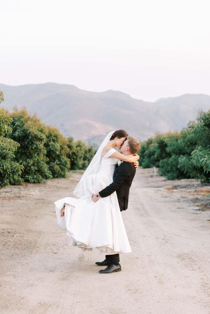 A groom in a black suit lifts and embraces his bride, who wears a white dress and veil, on a dirt path lined with green trees, with mountains visible in the background. Copyright - Lisa Leanne Photography, California