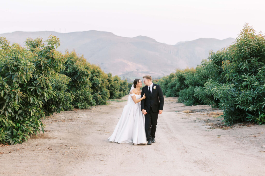 A bride in a white dress and a groom in a black suit walk hand in hand on a dirt path between green orchards, with mountains in the background under a soft sky. Copyright - Lisa Leanne Photography, California