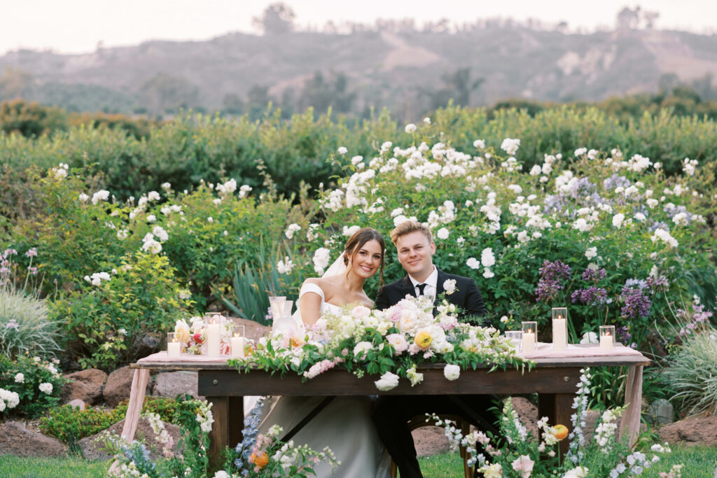 A bride and groom sit together, smiling, at a flower-adorned wooden table outdoors, surrounded by lush greenery and blooming flowers, with hills visible in the background. Copyright - Lisa Leanne Photography, California