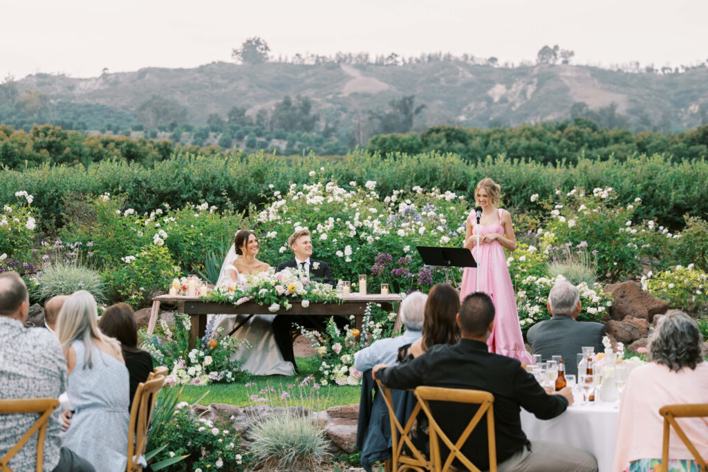 A woman in a pink dress gives a speech at an outdoor wedding reception. The bride and groom sit at a flower-adorned table, surrounded by guests, with lush greenery and hills in the background. Copyright - Lisa Leanne Photography, California
