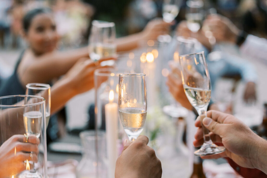People raise champagne glasses in a celebratory toast around a candlelit table, creating an atmosphere of joy and togetherness. The background is softly blurred, highlighting the festive mood. Copyright - Lisa Leanne Photography, California