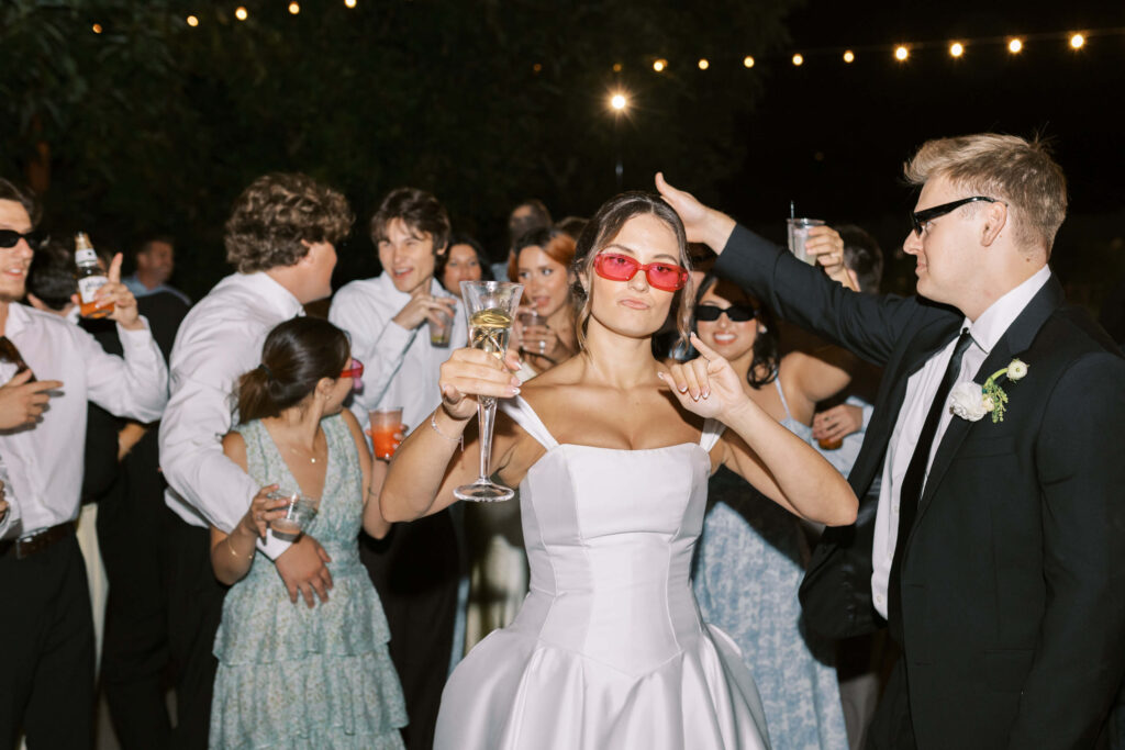 A bride in a white strapless dress and red sunglasses poses with a champagne glass, surrounded by laughing guests in formal attire at an outdoor nighttime celebration with string lights overhead. Copyright - Lisa Leanne Photography, California