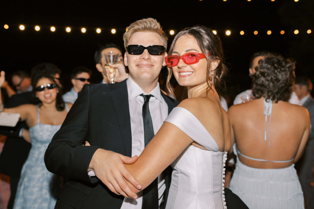 A couple wearing sunglasses dances together at a formal event, smiling at the camera. The woman is in an off-shoulder white dress, and the man is in a suit. People in formal attire are in the background under string lights. Copyright - Lisa Leanne Photography, California